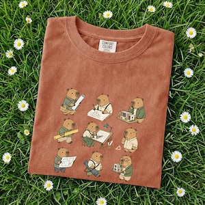 Könnte beinhalten: Ein terrakottafarbenes T-Shirt mit einem sich wiederholenden Muster von als Architekten gekleideten Wasserschweinen. Die Wasserschweine sind mit Bauplänen, Linealen und Miniatur-Gebäudemodellen dargestellt. Das Shirt hat einen Rundhalsausschnitt und das Label der Marke Comfort Colors.