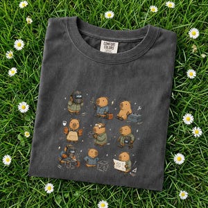 May include: Dark gray Comfort Colors t-shirt featuring a whimsical illustration of capybaras in various professions. The capybaras are depicted as a welder, carpenter, and engineer, among others. The shirt is laid on a bed of green grass with white daisies.