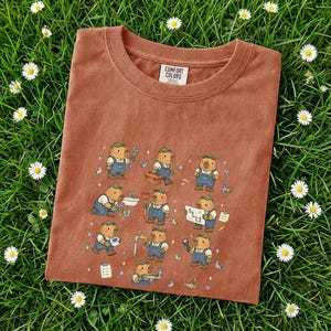 May include: A terracotta-coloured Comfort Colors t-shirt with a repeating pattern of cartoon capybaras dressed as plumbers. The capybaras wear blue overalls and green caps, holding tools and working on plumbing fixtures.