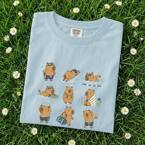 May include: A light blue Comfort Colors t-shirt featuring a playful graphic of capybaras engaged in various swimming activities. The capybaras are depicted wearing swim goggles, swimsuits, and holding medals. The shirt is laid on a bed of green grass with white daisies.