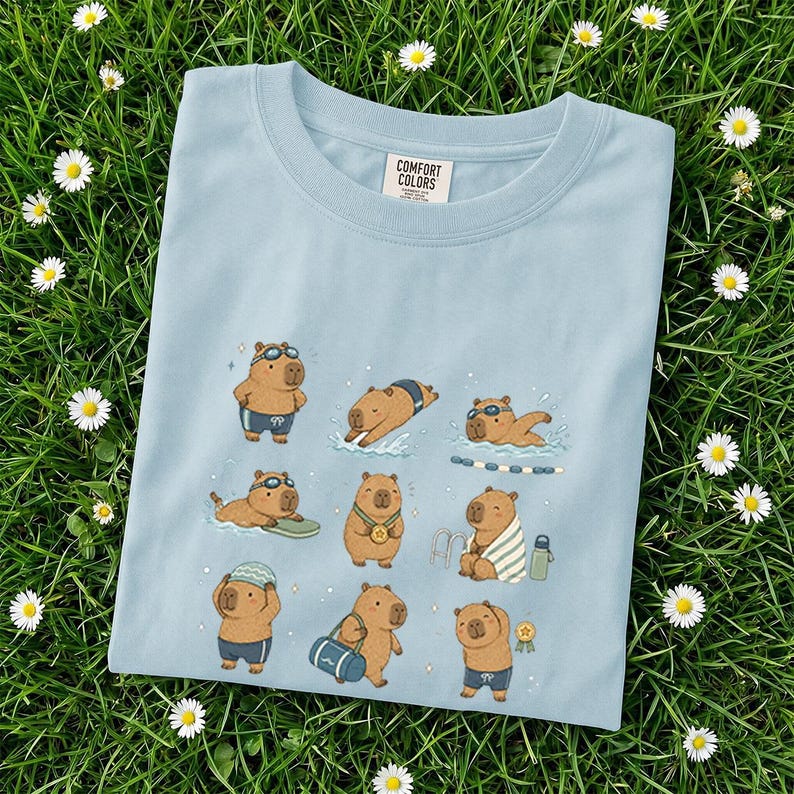 May include: A light blue Comfort Colors t-shirt featuring a playful graphic of capybaras engaged in various swimming activities. The capybaras are depicted wearing swim goggles, swimsuits, and holding medals. The shirt is laid on a bed of green grass with white daisies.