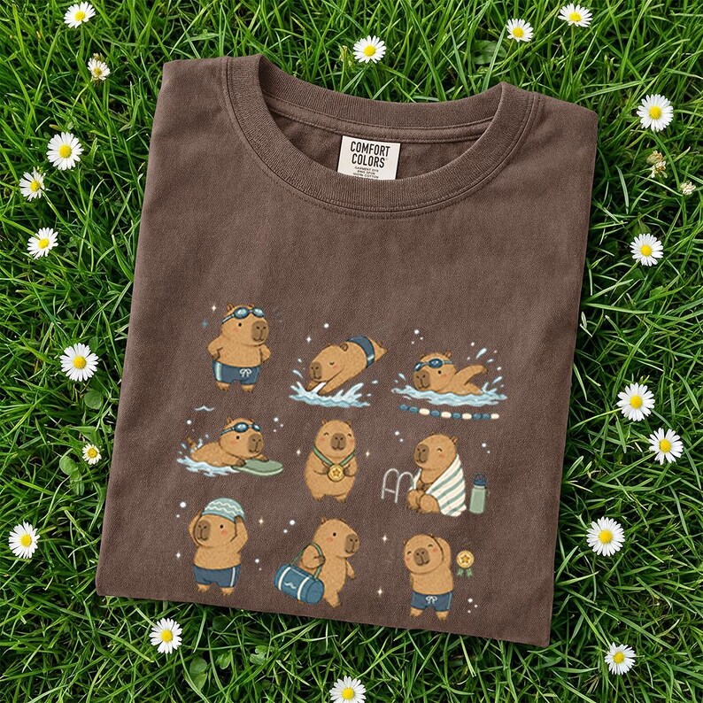 May include: Brown Comfort Colors t-shirt featuring a whimsical illustration of capybaras swimming and enjoying water activities. The capybaras are depicted in various poses, wearing swimsuits, goggles, and medals. The shirt is on a bed of green grass with white daisies.