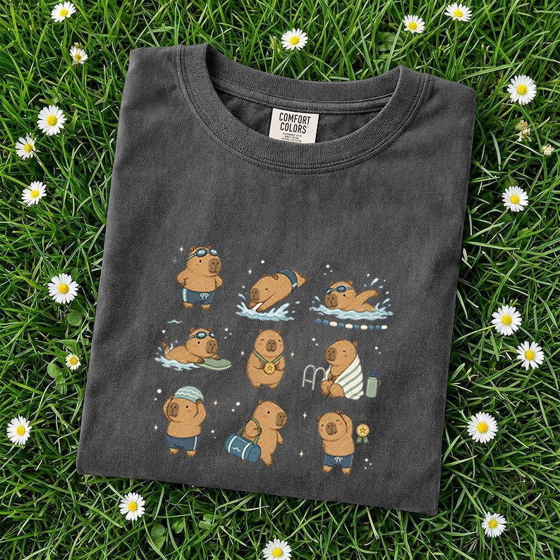 May include: Dark gray Comfort Colors t-shirt featuring a playful graphic of capybaras engaged in various swimming activities. The capybaras are depicted wearing swim goggles, swimsuits, and towels. The shirt is laid on a bed of green grass with small white flowers.