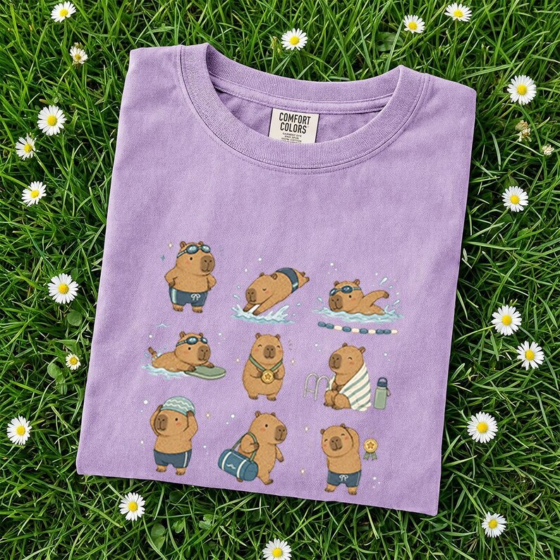 May include: A lilac Comfort Colors t-shirt featuring a playful illustration of capybaras enjoying various water activities. The capybaras are depicted swimming, sunbathing, and wearing goggles, swimsuits, and medals. The shirt is on a bed of green grass with white daisies.