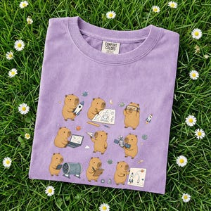 May include: A lilac Comfort Colors t-shirt featuring a whimsical illustration of capybaras engaged in space-themed activities. The design includes rockets, planets, and capybaras with goggles and notebooks. The shirt is laid on a bed of green grass with small white flowers.