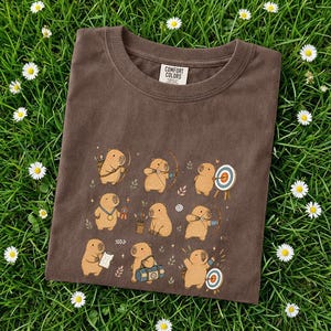 May include: Brown Comfort Colors t-shirt featuring a repeating pattern of cartoon capybaras engaged in archery and other activities. The shirt has a crew neck and the Comfort Colors logo is visible.