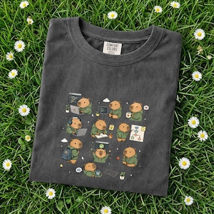 May include: Dark gray Comfort Colors t-shirt featuring a repeating graphic of cartoon capybaras in various tech-related poses. The capybaras are wearing green hoodies and interacting with laptops, coffee cups, and flowcharts. The shirt is laid on a bed of green grass with white daisies.