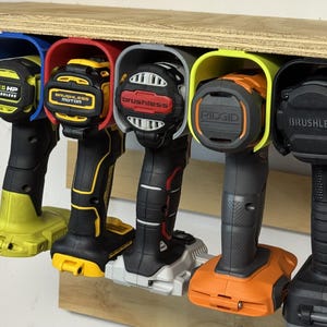 May include: A collection of cordless power drills in a variety of colors, including yellow, black, and orange, are mounted on a wooden shelf. Each drill is secured in a colored holder, with labels such as "ONE HP" and "BRUSHLESS" visible.
