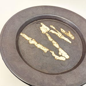 Puede incluir: Un plato de cerámica gris oscuro con un borde ancho. El centro del plato presenta un diseño abstracto texturizado en blanco roto y beige. El plato es redondo y parece hecho a mano.