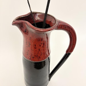 May include: A handmade ceramic pitcher with a red speckled top and a black base. The pitcher has a curved handle and a spout for pouring. Two dark branches are inside the pitcher. The pitcher is a decorative item.