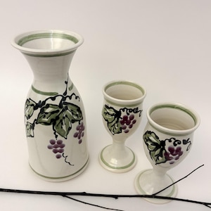 Może przedstawiać: Biały ceramiczny dzbanek i dwa pasujące kielichy na nóżkach, każdy ozdobiony ręcznie malowanymi zielonymi liśćmi winorośli i kiściami fioletowych winogron. Zestaw posiada proste zielone paski wokół krawędzi i podstaw.