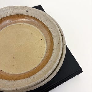 Puede incluir: Dos platos de cerámica en tonos beige y marrón. Los platos tienen una superficie moteada y texturizada, con un diseño circular. Están apilados sobre una superficie negra, sobre un fondo blanco.