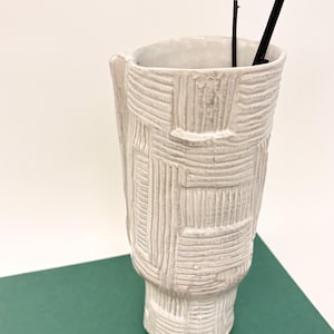 May include: A textured, off-white ceramic vase with a unique, geometric pattern. The vase has a rectangular shape with a flared base and a slightly curved top. Two dark, slender branches are placed inside the vase. The vase is set against a green and white background.