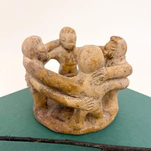 May include: A tan sculpture of five figures holding hands in a circle. The figures are stylized and appear to be made of a clay-like material. The sculpture is on a green surface.