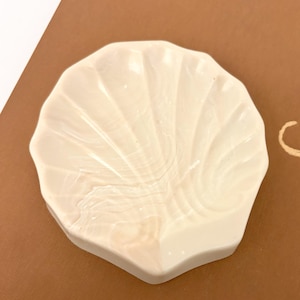 May include: A small, off-white, shell-shaped dish with a glossy finish. The surface features subtle, swirling marbled patterns in a lighter cream tone, mimicking the natural ridges and organic texture of a seashell in a compact, circular form.