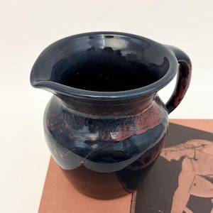 May include: A handcrafted ceramic pitcher with a dark blue glaze and a reddish-brown band around the middle. The pitcher has a curved handle and a spout for easy pouring. The pottery is likely handmade.