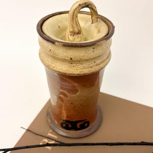 May include: A cylindrical ceramic vessel with a glossy, warm-toned brown glaze and a speckled cream-colored rim. The lid features a curved, handle-like protrusion. A black, stylized graphic symbol is visible near the base of the container.