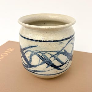 Op de afbeelding: Een crèmekleurige keramische pot met een donkerblauw abstract ontwerp. De pot heeft een afgeronde vorm met een licht uitlopende rand. Het blauwe ontwerp bestaat uit elkaar kruisende lijnen en wervelingen. De pot staat op een boek.