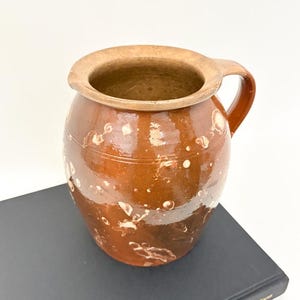May include: A brown ceramic jug with a handle and a wide opening. The jug has a speckled design with white spots and a light brown rim. The jug is sitting on a dark book.