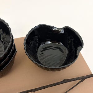 May include: A set of glossy black ceramic bowls with an irregular, organic rim shape and a vertically ribbed exterior texture. The bowls feature a deep, reflective interior surface and a scalloped edge design, presented in a stack of two.