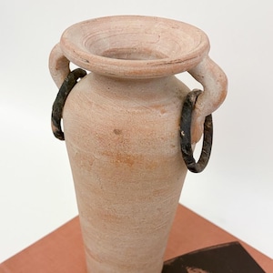 May include: A terracotta vase with a wide, shallow top and two curved handles. Dark metal rings are attached to the handles. The vase has a textured surface and a cylindrical body. The vase is sitting on a book.