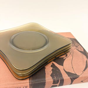 May include: Four square, smoky-grey glass coasters with a central circular indentation, stacked on a vintage book. The book has the title "Masterpieces of Greek Drawing and Painting" printed on the cover.
