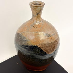 May include: A handcrafted ceramic vase with a rounded body and a narrow neck. The vase features a color palette of brown, tan, black, and orange, with a textured, layered design. The vase is a decorative item.