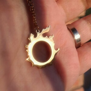 Puede incluir: Un collar con colgante dorado con un dise&ntilde;o similar al sol, un centro circular y bordes dentados. El colgante cuelga de una delicada cadena. La joya se exhibe sobre un fondo neutro.