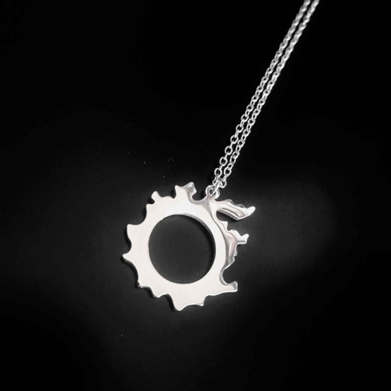 Puede incluir: Un collar con colgante plateado con un dise&ntilde;o calado distintivo. El colgante tiene un centro circular y un borde exterior irregular. Cuelga de una fina cadena, sobre un fondo oscuro.