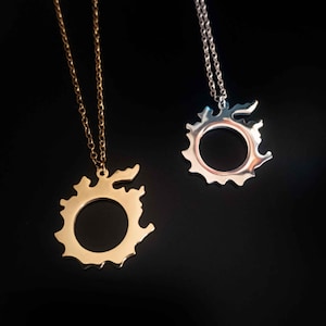 Puede incluir: Dos collares con colgantes de dise&ntilde;o circular y borde exterior dentado. Uno es dorado, el otro plateado. Ambos cuelgan de finas cadenas sobre un fondo negro.