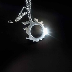 Puede incluir: Un collar con colgante plateado con una forma irregular y un c&iacute;rculo vac&iacute;o en el centro. El colgante cuelga de una cadena fina. El collar se muestra sobre un fondo oscuro.