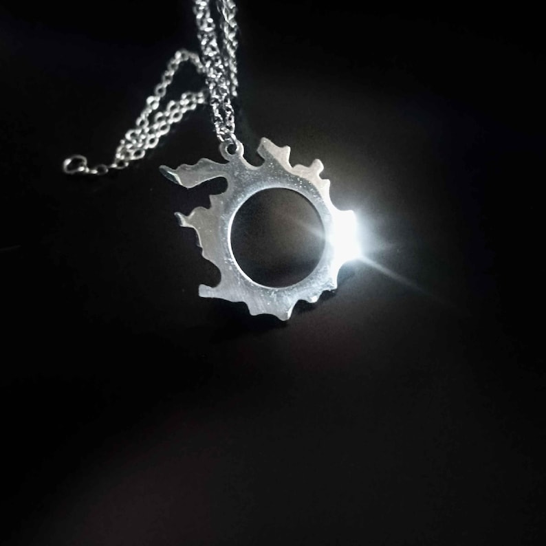 Puede incluir: Un collar con colgante plateado con una forma irregular y un c&iacute;rculo vac&iacute;o en el centro. El colgante cuelga de una cadena fina. El collar se muestra sobre un fondo oscuro.