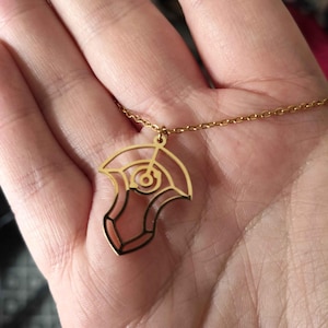 Puede incluir: Un collar con colgante dorado con un diseño calado estilizado. El colgante tiene una forma geométrica con un elemento circular central y está suspendido de una delicada cadena. El collar se muestra sobre un fondo neutro.