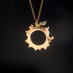 Puede incluir: Collar con colgante dorado con un dise&ntilde;o circular estilizado con centro abierto y bordes decorativos. El colgante cuelga de una delicada cadena dorada sobre un fondo oscuro. Es una joya.