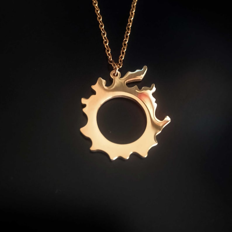 Puede incluir: Collar con colgante dorado con un dise&ntilde;o circular estilizado con centro abierto y bordes decorativos. El colgante cuelga de una delicada cadena dorada sobre un fondo oscuro. Es una joya.