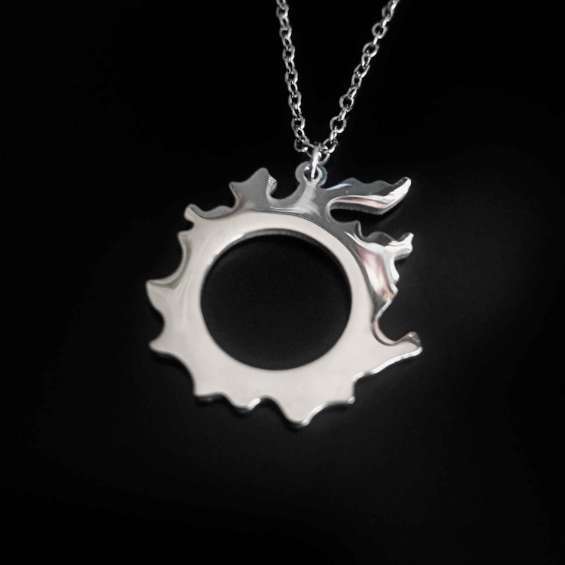 Puede incluir: Un collar con colgante plateado con un dise&ntilde;o distintivo, que presenta un c&iacute;rculo central abierto rodeado por un contorno irregular y puntiagudo. El colgante cuelga de una fina cadena, sobre un fondo negro. El dise&ntilde;o se asemeja a un sol o un emblema estilizado.