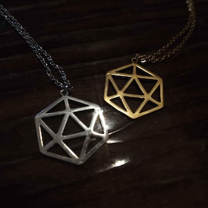 Peut inclure: Deux colliers avec des pendentifs géométriques en forme d'icosaèdre à cadre ouvert. Un pendentif est de couleur argentée et l'autre de couleur dorée. Chaque pendentif est suspendu à une fine chaîne en métal assortie, au design squelettique.