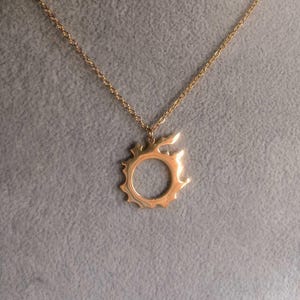 Puede incluir: Un collar con colgante dorado con un dise&ntilde;o de sol. El colgante tiene un centro circular con rayos puntiagudos que se extienden hacia afuera. La cadena del collar tambi&eacute;n es dorada, creando un aspecto cohesivo y elegante.