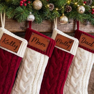 Puede incluir: Cuatro calcetines de Navidad con un patrón de punto trenzado en colores rojo y blanco alternos. Cada calcetín tiene un puño acanalado blanco con un parche de cuero marrón rectangular grabado con los nombres Kelly, Mom, Molly y Dad.