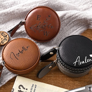 May include: Three round, leatherette cases in brown and black, each with a zip closure and a small leather pull tab. One case has the initials "E W" with a floral design, another has the name "Isabella", and the third has the name "Avalon" with a heart symbol.