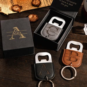 Könnte beinhalten: Drei personalisierte Flaschenöffner-Schlüsselanhänger aus grauem, schwarzem und braunem lederartigem Material mit Metallbeschlägen. Jeder zeigt einen großen Anfangsbuchstaben und einen Namen. Einer liegt in einer schwarzen Geschenkbox mit dem Aufdruck Alex.