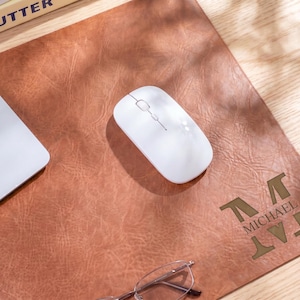 May include: A rectangular brown leather desk mat with a textured surface, featuring a white wireless computer mouse placed in the center and the name MICHAEL engraved in a dark font in the bottom right corner with geometric accents.