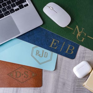 May include: A collection of four rectangular desk mats in green, blue, light blue, and brown, each featuring a unique gold-colored monogram or initial design. The mats are arranged near a laptop, a white computer mouse, and white wireless earbuds.