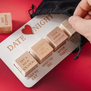 Puede incluir: Un juego de mesa para citas nocturnas de madera, con una base de madera blanca y dados de madera. La base tiene las palabras "DATE NIGHT" y un corazón rojo. Los dados tienen frases como "Cook Dinner Together" y "Sexy Time".
