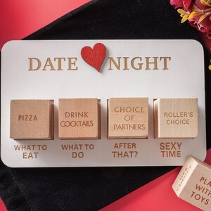 Może przedstawiać: Biała drewniana gra Date Night z czerwonym sercem i grawerowanym tekstem. Zawiera drewniane klocki z opcjami takimi jak "Pizza" i "Drink Cocktails". Obecna jest również kostka z napisem "Play with Toys".
