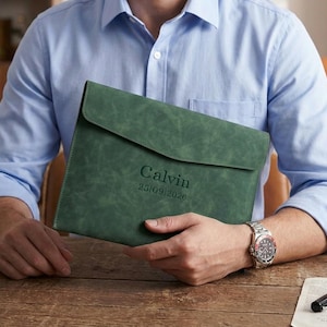 Könnte beinhalten: Eine dunkelgrüne Dokumentenmappe aus Leder mit Klappenverschluss. Der Name "Calvin" und das Datum "25/09/2026" sind auf der Vorderseite eingeprägt. Der Halter wird von einer Person gehalten, die ein hellblaues Hemd trägt.