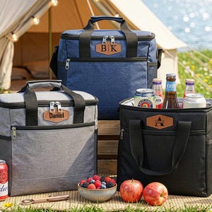 May include: Three personalized cooler bags in gray, blue, and black, with black handles and zippers. The bags have leatherette name tags. The scene includes drinks, fruit, and a tent in the background, suggesting a picnic or outdoor event.
