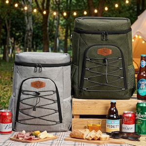 May include: Two insulated cooler backpacks, one gray and one olive green, are displayed outdoors. Each has a front pocket with a bungee cord design and a leather patch. Various bottles and cans of beer, along with snacks, are arranged on a picnic blanket.