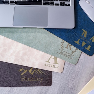 Könnte beinhalten: Eine Sammlung rechteckiger Schreibtischunterlagen in Dunkelblau, Salbeigrün, hellem Beige und Dunkelbraun, jeweils mit personalisiertem goldfarbenem Text und Grafiken wie den Namen Michael, Robert, Arthur und Stanley, platziert neben einem Laptop.