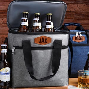 Op de afbeelding: Een grijze koeltas met zwarte handvatten en een bruine kunstlederen patch met de tekst JAC. De tas staat open en bevat drie glazen bierflesjes. Op de achtergrond is een tweede blauwe koeltas met een patch met de tekst David zichtbaar.